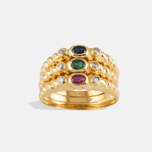 May include: A gold ring featuring three stacked bands with a twisted rope texture. The center of each band holds an oval gemstone: a dark blue sapphire, a green emerald, and a red ruby, each flanked by small round clear diamonds.
