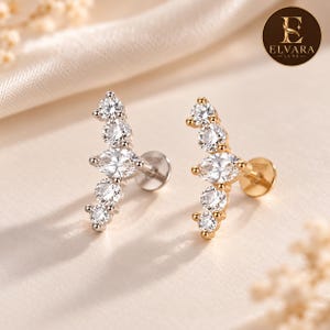 May include: Two stud earrings with flat back posts, one in silver tone and one in gold tone. Each earring features a curved design set with four round brilliant-cut stones and one central marquise-cut stone. The logo ELVARA LUXE is visible.