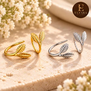 May include: Two pairs of ear cuffs, one gold-toned and one silver-toned, each featuring a dual-leaf design. The jewelry is displayed alongside white flowers, with the Elvara Luxe logo visible in the upper right corner of the image.