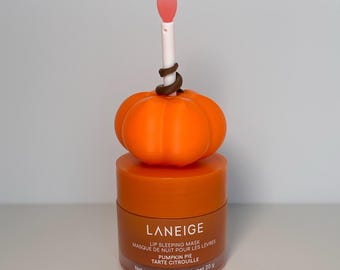 Pode incluir: Máscara labial noturna Laneige com aroma de Pumpkin Pie, peso líquido 20 g. O pote laranja tem uma tampa com um ornamento de abóbora laranja brilhante e um pequeno aplicador branco com ponta rosa saindo do centro.