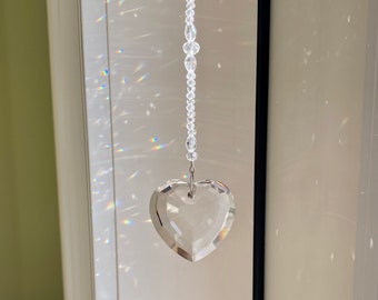Op de afbeelding: Een heldere, facetgeslepen kristallen hartvormige hanger die aan een ketting van heldere kralen hangt. Het hart is bevestigd met een zilverkleurige metalen lus. Licht breekt door het kristal en creëert regenboogpatronen.