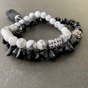 May include: A black and white beaded bracelet with a silver charm and a black tassel. The bracelet is made of small, irregularly shaped beads.