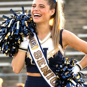 May include: A navy blue and white cheerleading uniform featuring a sleeveless crop top with gold star accents and a matching pleated skirt. A white sash with gold glitter text reading CHEER SENIOR is worn across the chest, paired with navy and gold pom-poms.