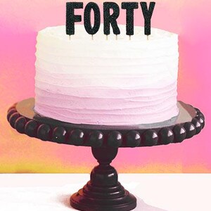 May include: A white and pink ombre cake on a black beaded cake stand. The cake is topped with black glitter "FORTY" cake toppers. The background is a gradient of pink and yellow.