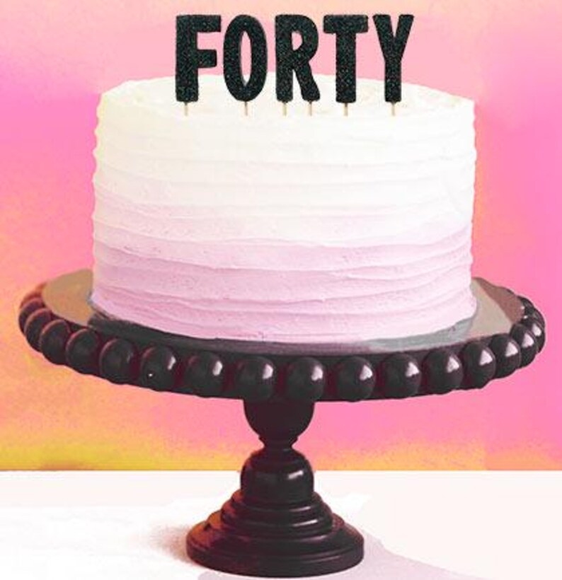 May include: A white and pink ombre cake on a black beaded cake stand. The cake is topped with black glitter "FORTY" cake toppers. The background is a gradient of pink and yellow.