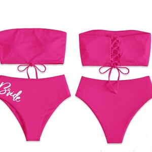 May include: A pink strapless bikini top with a lace-up back and matching high-waisted bikini bottoms. The bottoms have the word "Bride" written in white on the front.