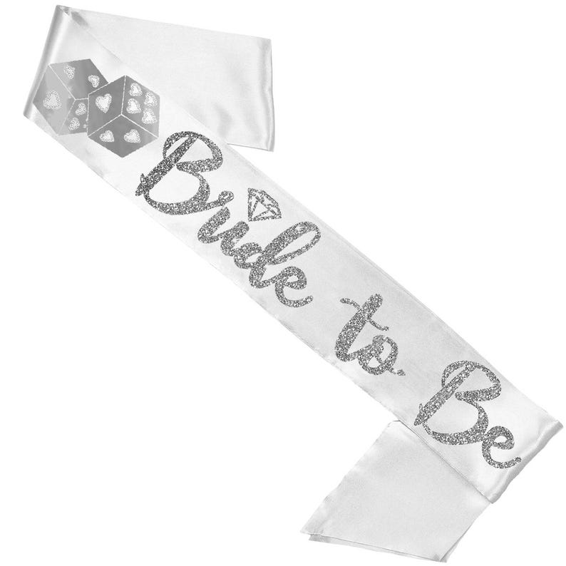 以下が含まれることがあります： 「Bride to Be」と銀色のグリッターテキストが入った白いサテンサッシュ。サッシュにはダイヤモンドのグラフィックとハートのシンボルが付いた2つのサイコロがあります。ブライダルシャワーや独身最後のパーティーに最適なアクセサリーです。
