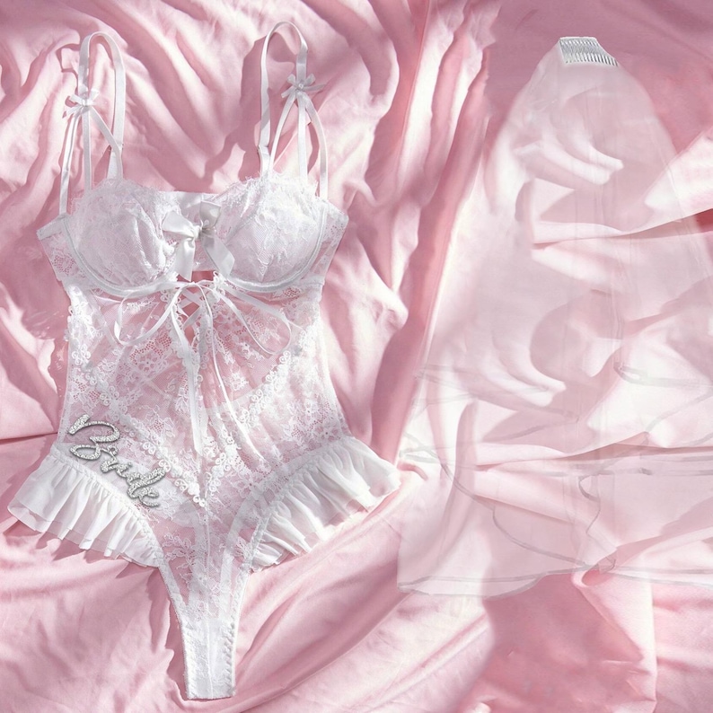 K&ouml;nnte beinhalten: Wei&szlig;e Spitzenunterw&auml;sche mit R&uuml;schenbesatz und dem aufgen&auml;hten Wort "Bride" auf der Vorderseite. Die W&auml;sche liegt auf einem rosa Satin-Stoff.