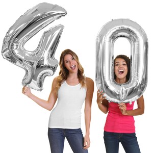 May include: Two women holding large, silver foil balloons shaped as the numbers 4 and 0, forming the number 40. The women are smiling and wearing casual clothing, a white tank top and a pink tank top with blue jeans. The balloons are against a white background.