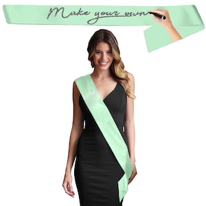 May include: A mint green sash with the text "Make your own" written in glitter. A person is wearing a mint green sash over a black dress. The sash is blank and ready to be customized. The image is on a white background.