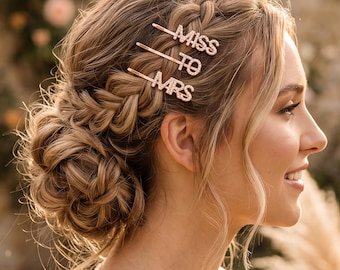 May include: Three rose gold hair clips featuring clear rhinestones that spell out the words MISS TO MRS. The clips are secured into a braided hairstyle, with each individual pin displaying one word of the phrase in a clear, capital letter font.
