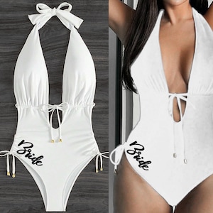 May include: A white halter-neck swimsuit with a plunging neckline and the word "Bride" printed in black script. The swimsuit features tie details at the sides and a bow at the neck, perfect for a bachelorette party or bridal event.