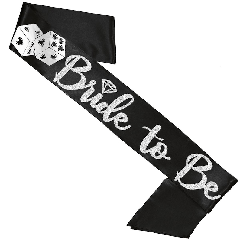 以下が含まれることがあります： 「Bride to Be」の文字とダイヤモンドのグラフィックが施された、黒いサテンのサッシュ。左上にはハートとドットの模様が入った白いサイコロが2つあります。ブライダルのお祝いにぴったりの楽しいアクセサリーです。