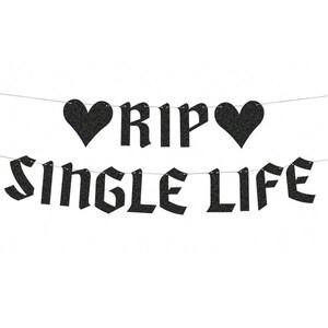 May include: A black glittery banner featuring two heart shapes and the text RIP SINGLE LIFE in a gothic-style font. The letters and hearts are strung together on a thin silver string, creating a two-line decorative party garland.