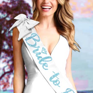 May include: White satin sash with the text "Bride to Be" in light blue glitter script. The sash features a diamond graphic and a large bow at the shoulder. The sash is worn over a white dress.