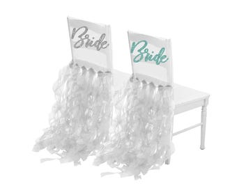 May include: Two white chairs with sheer, ruffled fabric draped over the backs. The word "Bride" is written in glittery script on the chair backs, one in silver and the other in teal. The chairs are on a white bench.