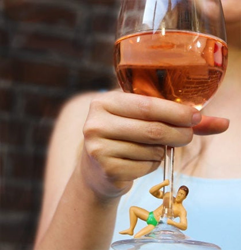 Puede incluir: Una copa de vino rosado sostenida por una persona. El tallo de la copa presenta una peque&ntilde;a figura de un hombre en traje de ba&ntilde;o verde. La copa de vino est&aacute; llena de un l&iacute;quido rosa claro.