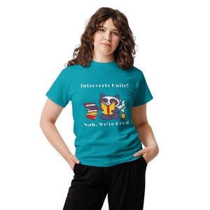 Puede incluir: Una camiseta de manga corta color verde azulado con un gráfico de un panda leyendo un libro junto a una pila de libros y una taza humeante. El texto "Introverts Unite!" aparece arriba y "Nah, We're Good" está impreso bajo el gráfico.