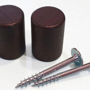 Puede incluir: Dos tapas cilíndricas de madera de color marrón oscuro con partes superiores planas y dos tornillos de metal de color plateado con cabezas planas y vástagos roscados. Los componentes de madera son sólidos y opacos, mientras que los tornillos tienen un acabado metálico.