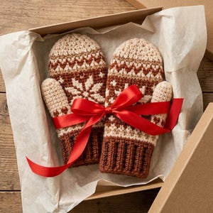 Könnte beinhalten: Ein Paar gehäkelte Fäustlinge mit einem Fair-Isle-Muster in Creme-, Beige- und Dunkelbrauntönen. Die Fäustlinge haben ein geripptes Bündchen und sind mit einem leuchtend roten Satinband zusammengebunden, präsentiert in einer Pappschachtel mit Seidenpapier.