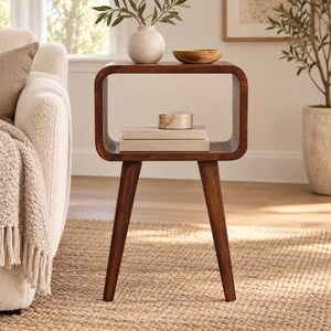 May include: A dark brown wooden side table with a rectangular open frame design and four tapered legs. The table surface holds a round off-white vase, a small wooden bowl, a circular stone container, and two stacked rectangular books.