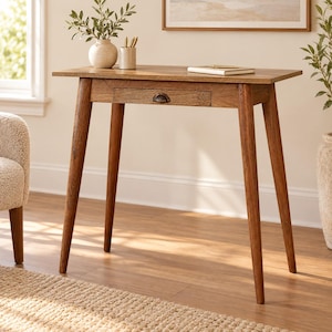 May include: A wooden writing desk with a rectangular top, four tapered legs, and a single central drawer featuring a small, dark metal cup pull handle. The desk has a warm, medium-brown wood grain finish and a clean, simple structural design.
