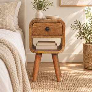 May include: A light brown wooden nightstand featuring a single drawer with a dark circular knob, an open storage shelf containing two stacked books, and four tapered legs. The piece has rounded corners and a smooth, natural wood grain finish.