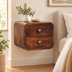 May include: A wall-mounted, dark brown wooden nightstand with rounded corners and two drawers. Each drawer features a single, small, round metallic knob. A white ceramic vase with green foliage sits on top of the nightstand.