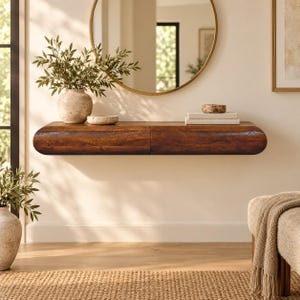 May include: A dark brown wooden floating console table with rounded edges, mounted on a wall. The surface holds a beige ceramic vase with green foliage, a small stone bowl, and two stacked white books beneath a round wall mirror.