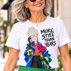 Puede incluir: Una camiseta blanca de manga corta con una ilustración gráfica de una persona con el pelo gris, gafas, una blusa roja, guantes negros y una chaqueta con estampado azul, amarillo y negro con una flor rosa. El texto dice MORE STYLE THAN YEARS.