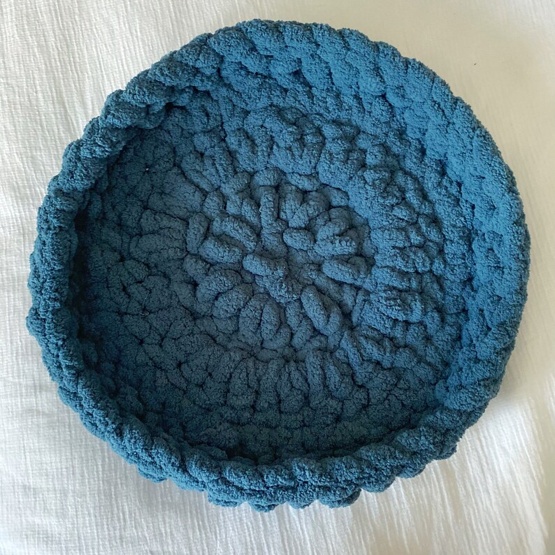 May include: A large, round, blue crocheted pet bed. The bed is made of thick, soft yarn and has a deep, comfortable center.