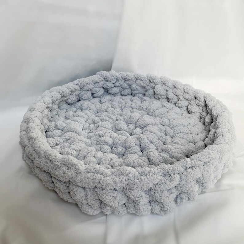 May include: A light gray round pet bed made of crocheted yarn. The bed is made of soft, fluffy yarn and has a raised edge.