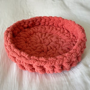 May include: A coral-coloured knitted pet bed made of soft, fluffy yarn. The bed is round and has a raised edge.