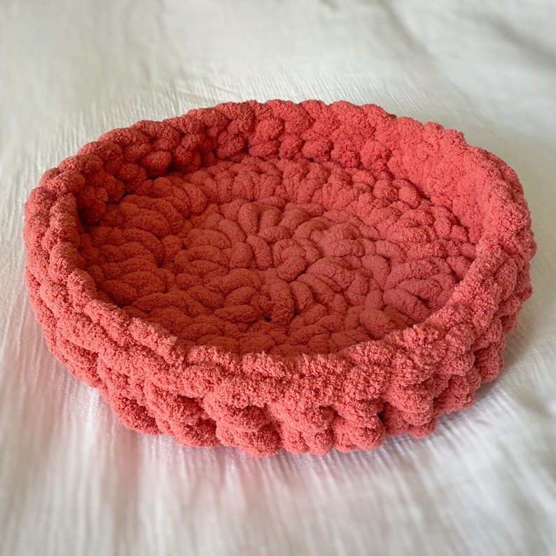 May include: A coral-coloured knitted pet bed made of soft, fluffy yarn. The bed is round and has a raised edge.