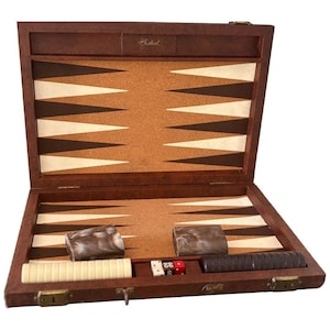 May include: A brown wooden backgammon board with a cork playing surface featuring dark brown and cream-colored triangles. The set includes two marbled dice cups, a row of cream checkers, a row of dark brown checkers, and two standard dice.