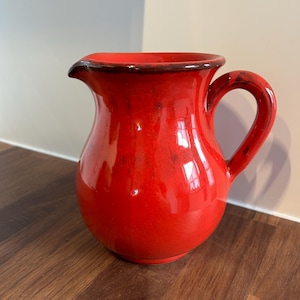 Può includere: Una brocca in ceramica rosso brillante con corpo arrotondato, manico curvo e beccuccio. Lo smalto presenta una finitura lucida con sottili sfumature rosso scuro e marrone attorno al bordo e al manico, creando un aspetto strutturato.