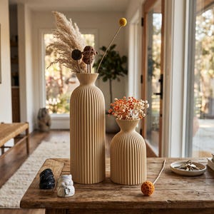 May include: Two beige, vertically ribbed ceramic vases sit on a wooden table. The taller, slender vase holds dried pampas grass and a yellow billy ball, while the shorter, rounded vase contains a bouquet of small orange and white flowers.