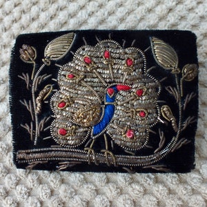 May include: A black velvet patch featuring a gold and silver embroidered peacock design. The peacock has a blue body and red accents. The patch is surrounded by a floral design.