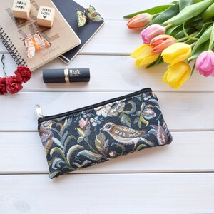 May include: A black zippered pouch with a floral and bird tapestry design. The pouch is on a white wooden surface with a lipstick, flowers, and a notebook in the background.