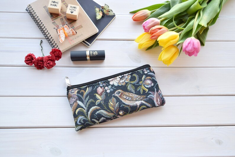 May include: A black zippered pouch with a floral and bird tapestry design. The pouch is on a white wooden surface with a lipstick, flowers, and a notebook in the background.