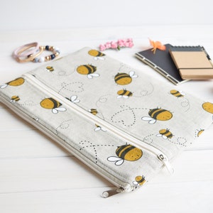 Könnte beinhalten: Eine beige Reißverschlusstasche mit einem weißen Reißverschluss und einem Muster aus gelben Bienen auf weißem Hintergrund. Die Tasche liegt auf einer weißen Oberfläche.