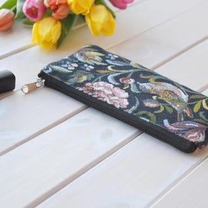 May include: A black zippered pouch with a floral and bird tapestry design. The pouch is lying on a white wooden surface.