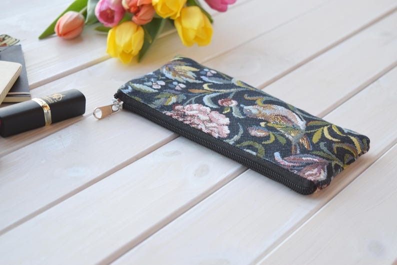 May include: A black zippered pouch with a floral and bird tapestry design. The pouch is lying on a white wooden surface.