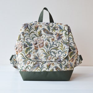 Può includere: Uno zaino color crema con un motivo floreale e di uccelli. Lo zaino ha una cinghia verde e un fondo verde.