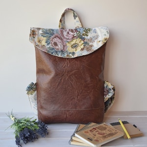 May include: A brown leather backpack with a floral fabric flap. The backpack has two side pockets and a zippered closure. The floral fabric is a mix of pink, blue, and yellow flowers on a white background.