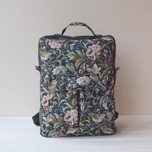 Puede incluir: Una mochila negra con un diseño de tapicería floral con flores rosas y blancas, hojas verdes y pájaros pequeños. La mochila tiene un asa superior y correas ajustables.