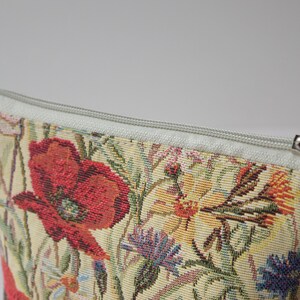 Pu&ograve; includere: Una pochette floreale con cerniera, con sfondo bianco e un motivo di papavero rosso. La pochette &egrave; realizzata in tessuto a tappezzeria e ha una chiusura a cerniera.