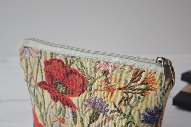 Pu&ograve; includere: Una pochette floreale con cerniera, con sfondo bianco e un motivo di papavero rosso. La pochette &egrave; realizzata in tessuto a tappezzeria e ha una chiusura a cerniera.