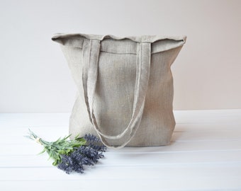 May include: A large, natural linen tote bag with two long handles. The bag is empty and sitting on a white surface. A small bouquet of lavender is in front of the bag.