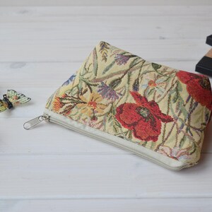 Pu&ograve; includere: Una pochette beige con cerniera con un motivo floreale che presenta papaveri rossi, fiori gialli e viola e foglie verdi. La pochette &egrave; realizzata in tessuto a tappezzeria.